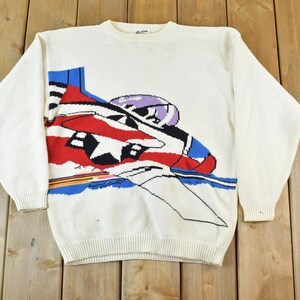 May include: A white knitted sweater with a colorful graphic of a plane flying over a blue and white striped background. The plane is red, white, and blue with a black star on the wing.