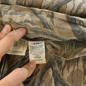 Op de afbeelding: Close-up van een stof met camouflagepatroon. De stof is een mix van bruine, groene en beige kleuren. Een label met wasvoorschriften en maat XXL is zichtbaar.