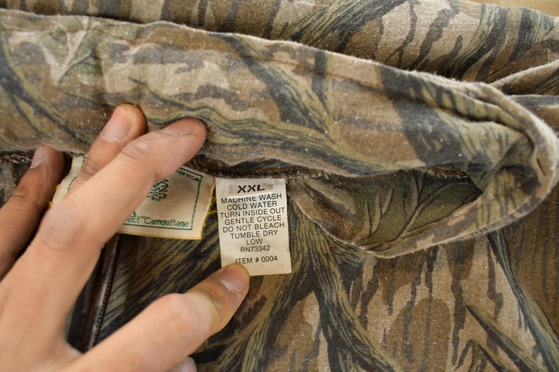 Op de afbeelding: Close-up van een stof met camouflagepatroon. De stof is een mix van bruine, groene en beige kleuren. Een label met wasvoorschriften en maat XXL is zichtbaar.