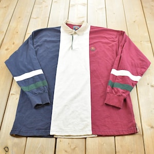May include: A long-sleeve rugby shirt featuring a color-blocked design with a navy blue left side, a white center panel, and a maroon right side. The shirt has a beige collar, a button placket, and sleeves with white and green horizontal stripes.