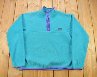 May include: A teal blue fleece pullover with a purple trim and a single button closure at the neck. The Patagonia logo is visible on the front of the sweater.