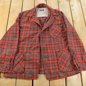 May include: A red and grey checked jacket with two front pockets and a chest pocket. The jacket features a collared neckline and button fastenings. The fabric has a classic, vintage appearance.