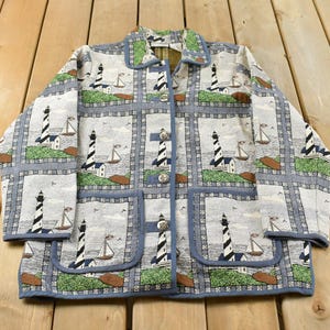 May include: A long-sleeve button-up jacket featuring a repeating grid pattern of lighthouses and sailboats. The fabric is light grey with blue, green, and brown accents. It includes a standard collar and two large square pockets on the front.