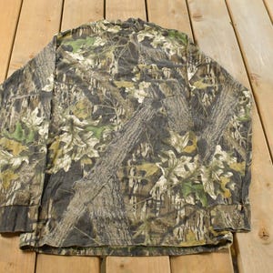 May include: Long-sleeved camouflage shirt in shades of brown, green, and tan. The pattern mimics tree bark and foliage, creating a naturalistic effect. The shirt is displayed flat on a wooden surface.