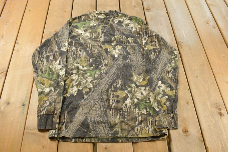 May include: Long-sleeved camouflage shirt in shades of brown, green, and tan. The pattern mimics tree bark and foliage, creating a naturalistic effect. The shirt is displayed flat on a wooden surface.