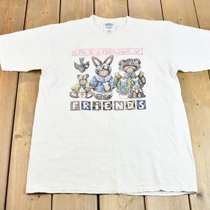 May include: White t-shirt featuring a graphic of a bunny, two bears, and a bird. The graphic is surrounded by a pink banner that reads "Life is a patchwork of" and a banner with the word "FRIENDS" in colourful blocks.