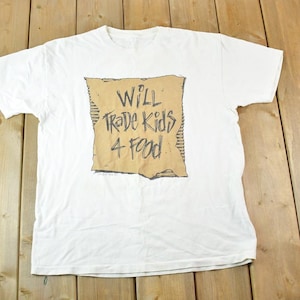 May include: White t-shirt featuring a distressed brown graphic with the text "Will Trade Kids 4 Food" in black. The shirt is displayed on a wooden surface.