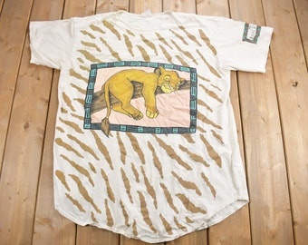 May include: White t-shirt with a brown and white zebra print. A cartoon lion cub is sleeping on a branch in a framed image on the front of the shirt. The shirt has a tag that says "Lion King".