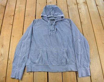 May include: A pale blue hooded sweatshirt with a kangaroo pocket and elbow patches. The sweatshirt features a drawstring neckline and a small logo on the chest. The garment is made of a soft, faded material.