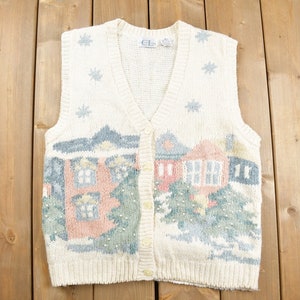 May include: A cream-colored knitted vest with a winter scene design. The vest has a V-neck and button closure. The design features houses, trees, and snowflakes in shades of pink, blue, and green.
