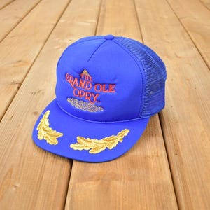 Puede incluir: Gorra de camionero azul con el texto "The Grand Ole Opry" en bordado rojo y dorado. La gorra presenta una parte trasera de malla y detalles de hojas doradas en la visera, sobre una superficie de madera.