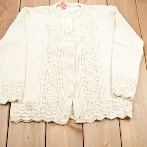 Puede incluir: Un cárdigan blanco de ganchillo con cierre de botones y bordes festoneados.