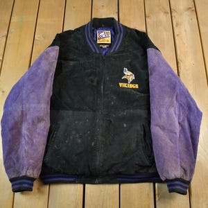 May include: Black and purple vintage jacket with the Minnesota Vikings logo. The jacket has a black body with purple suede sleeves and a blue and black striped collar and cuffs. The Vikings logo is embroidered in gold.