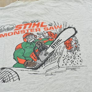 Op de afbeelding: Een lichtgrijs t-shirt met een afbeelding van een stripfiguur die beschermende kleding draagt en een Stihl-kettingzaag bedient. De tekst STIHL MONSTER SAW staat in oranje boven het figuur, met houtsnippers die van het zaagblad vliegen.