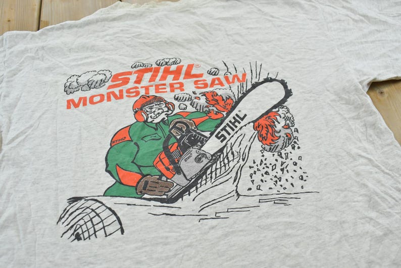 Op de afbeelding: Een lichtgrijs t-shirt met een afbeelding van een stripfiguur die beschermende kleding draagt en een Stihl-kettingzaag bedient. De tekst STIHL MONSTER SAW staat in oranje boven het figuur, met houtsnippers die van het zaagblad vliegen.