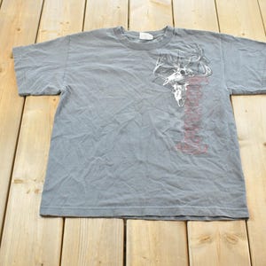 May include: Gray short-sleeved t-shirt with a graphic of a skull and antlers in white and red. The shirt is laid flat on a wooden surface. The t-shirt is a crew neck style.