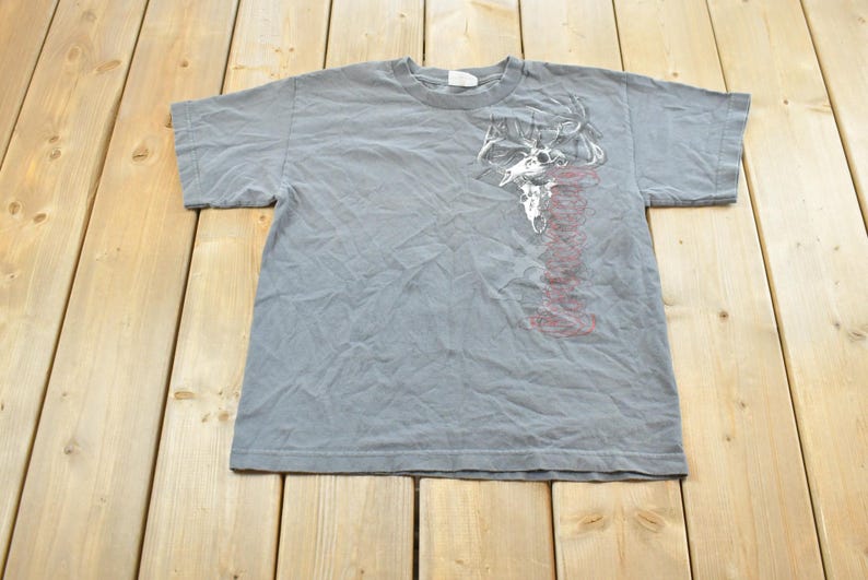 May include: Gray short-sleeved t-shirt with a graphic of a skull and antlers in white and red. The shirt is laid flat on a wooden surface. The t-shirt is a crew neck style.