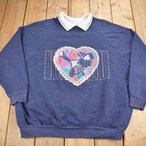 May include: A navy blue sweatshirt with a white collared neckline and a large embroidered heart on the front. The heart is made of colorful patchwork fabric and has a white lace trim.