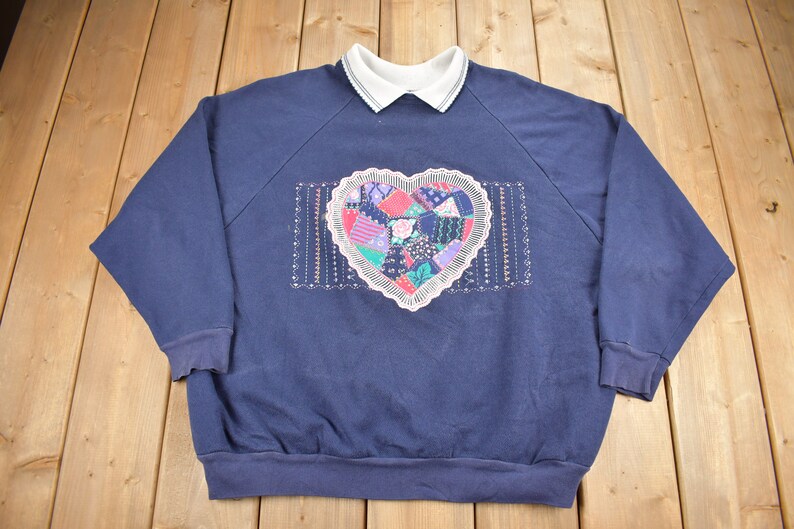 May include: A navy blue sweatshirt with a white collared neckline and a large embroidered heart on the front. The heart is made of colorful patchwork fabric and has a white lace trim.