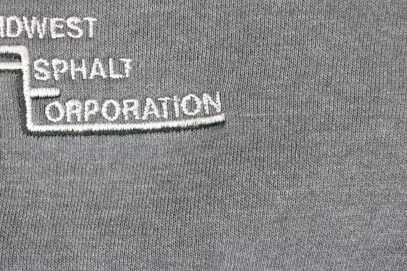 Puede incluir: Primer plano de una tela gris con texto bordado en blanco. El texto dice "MIDWEST ASPHALT CORPORATION" en una fuente estilizada. La tela tiene una apariencia texturizada.