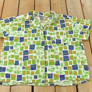 May include: Short-sleeved button-down shirt with a white background and a repeating pattern of green, blue, and orange squares, and palm tree silhouettes. The shirt has a classic collar and is made of a lightweight fabric.