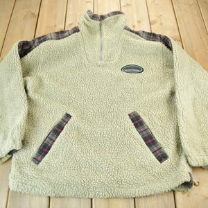 May include: A beige fleece pullover with a half-zip closure and plaid accents on the shoulders and pockets. The pullover has a small oval logo on the chest.