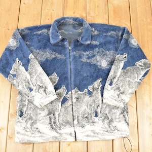 Könnte beinhalten: Blaue Jacke mit Reißverschluss und Wolfsmotiv. Die Jacke hat einen dunkelblauen Hintergrund mit einem weißen und grauen heulenden Wolfsmotiv. Die Jacke hat einen Kragen und einen durchgehenden Reißverschluss.