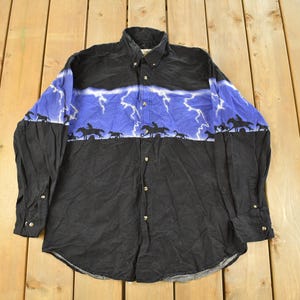 Puede incluir: Camisa negra con botones con un diseño de relámpagos azules y blancos en el pecho y los hombros. La camisa presenta una silueta de jinetes a caballo en la parte inferior de la sección azul. Una camisa de temática occidental.
