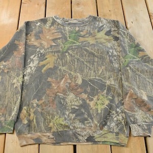 May include: Crew neck sweatshirt with a camouflage pattern featuring brown, green, and tan leaf and branch designs. The long-sleeved top is made of a soft material and is suitable for casual wear.