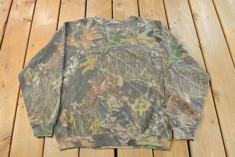 May include: Crew neck sweatshirt with a camouflage pattern featuring brown, green, and tan leaf and branch designs. The long-sleeved top is made of a soft material and is suitable for casual wear.