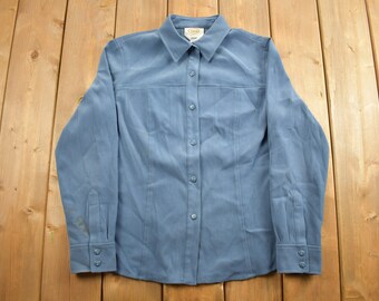 Può includere: Una camicia a maniche lunghe di colore azzurro chiaro con bottoni e colletto. La camicia ha una tasca sul petto e chiusura con bottoni.