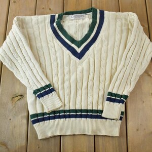 Può includere: Un maglione bianco a treccia con strisce verdi e blu navy intorno al collo e ai polsini. Il maglione ha un collo a V.