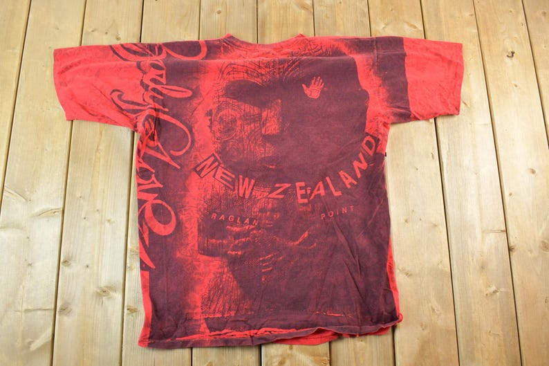 May include: Red t-shirt with a dark red graphic design featuring a face, handprint, and the words "Body Love" and "New Zealand Raglan Point". The shirt is laid flat on a wooden surface.