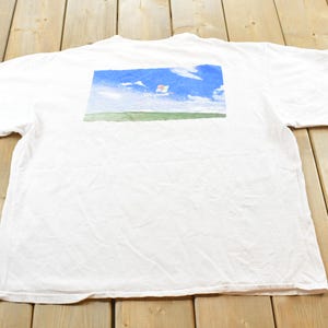 May include: White t-shirt with a rectangular graphic on the back featuring a blue sky with clouds and a green field. The graphic has a watercolor painting style. The shirt is made of a soft material.
