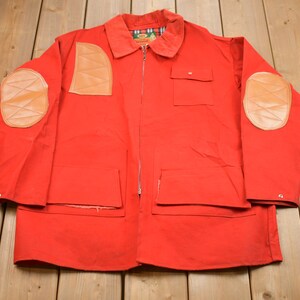 May include: A red jacket with leather elbow patches and a zipper closure. The jacket has two large pockets on the front and a smaller pocket on the left chest.
