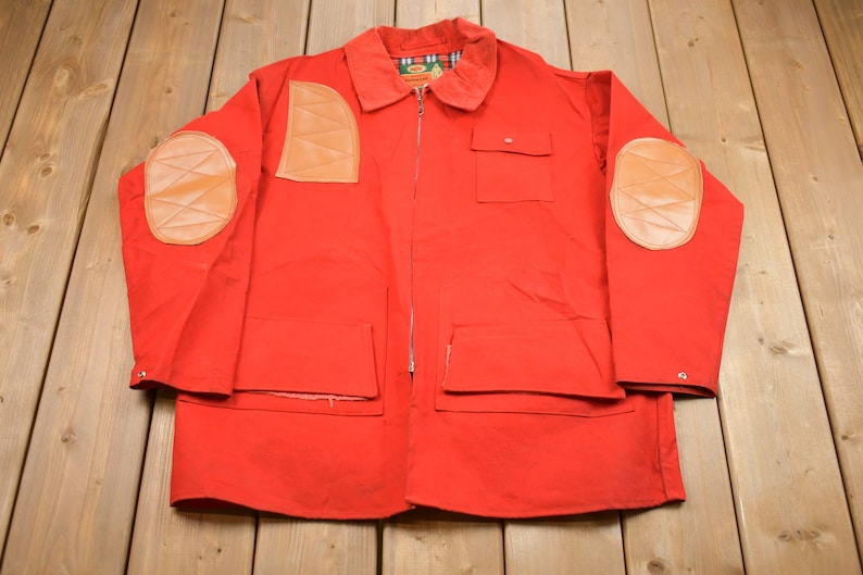 May include: A red jacket with leather elbow patches and a zipper closure. The jacket has two large pockets on the front and a smaller pocket on the left chest.