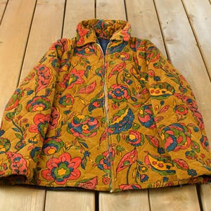 May include: A quilted jacket in a mustard yellow colour featuring a repeating floral pattern in shades of red, blue, and green. The jacket has a full-length front zip, a fold-over collar, and long sleeves with a diamond-stitched quilted texture.