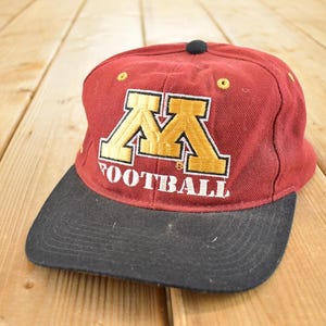May include: Maroon and black baseball cap featuring the word "FOOTBALL" and a gold and maroon "M" logo. The cap has a black brim and a small black button on top. The hat is on a wooden surface.