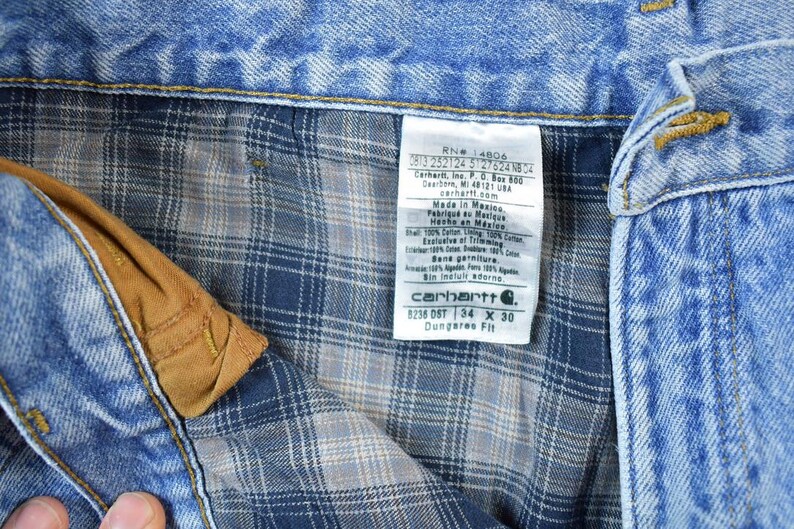 K&ouml;nnte beinhalten: Nahaufnahme einer blauen Jeans mit kariertem Futter. Die Jeans hat eine braune Tasche und ein Carhartt-Etikett. Das Etikett zeigt die Gr&ouml;&szlig;e 34 x 30 Zoll an. Die Artikelnummer ist 1236 DST.