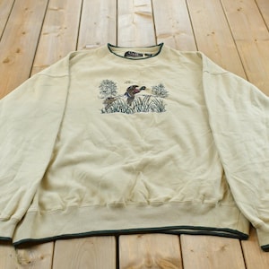 May include: A cream-coloured long-sleeve sweatshirt featuring a dark green trim at the crew neckline and cuffs. The centre of the chest displays an embroidered graphic of a duck flying over tall green grass and trees with the text C&B Sport.