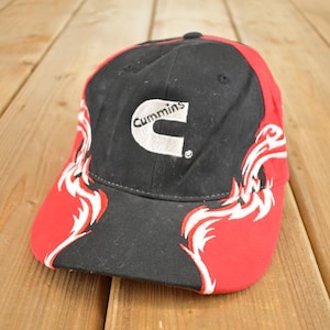Op de afbeelding: Zwarte en rode baseballcap met het witte Cummins-logo. De cap heeft een gebogen klep en een decoratief ontwerp in wit en rood aan de zijkanten.