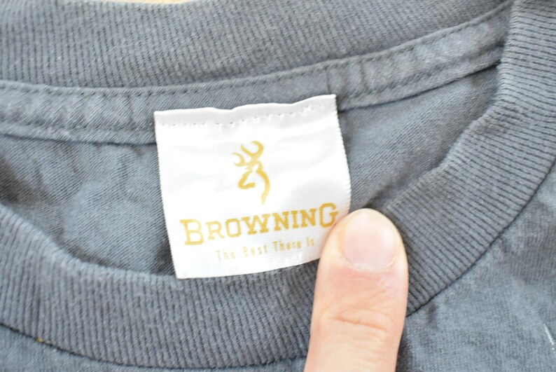 May include: Close-up of a gray t-shirt collar with a white tag. The tag features the word "BROWNING" in gold lettering, along with a gold deer logo and the text "The Best There Is".