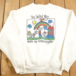 Puede incluir: Sudadera blanca con un estampado gráfico colorido de una casa, un arcoíris, un sol, un pájaro y personas. El texto "The United Way makes my rainbow brighter" está impreso debajo del gráfico.