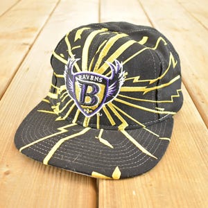 May include: Black and gold baseball cap featuring a stylized design with lightning bolts. The cap displays the Baltimore Ravens logo, a purple and white shield with a large "B" and the word "RAVENS".