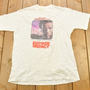 Pode incluir: Camiseta branca com um gráfico de Clint Eastwood como Harry Callahan no filme "Impacto Repentino". O gráfico apresenta um close-up do rosto de Eastwood com um fundo vermelho e roxo.