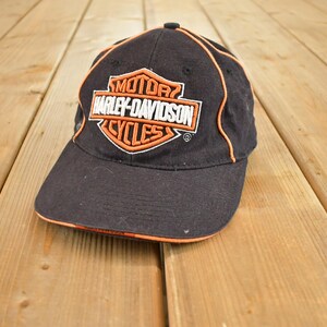 Peut inclure: Casquette de baseball noire avec le logo Harley-Davidson Cycles en orange et blanc. La casquette a une bordure orange. Le fond est en bois.