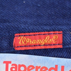 May include: A red label with the word "Wrangler" in gold lettering on a dark blue denim background.
