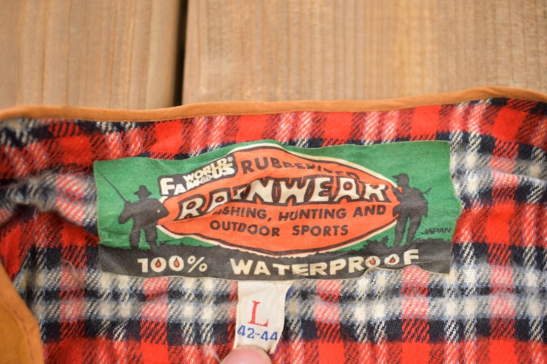 May include: A green and red plaid flannel shirt with a brown leather trim. The shirt has a label that reads "World's Famous Rubberized Rainwear Fishing, Hunting and Outdoor Sports 100% Waterproof" and a size tag that reads "L 42-44".