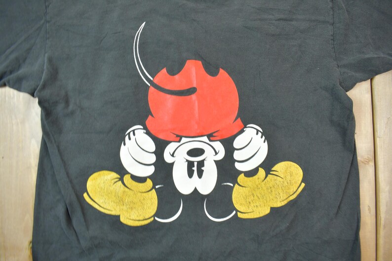May include: Black t-shirt with a cartoon image of Mickey Mouse wearing a red hat and looking down. Mickey Mouse is wearing yellow shoes and white gloves.
