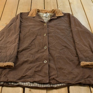 May include: A brown long-sleeve jacket featuring a tan corduroy collar, a button-down front closure, one chest pocket, and two large lower patch pockets. The interior is lined with a white and brown checkered fabric, and the sleeve cuffs are folded.